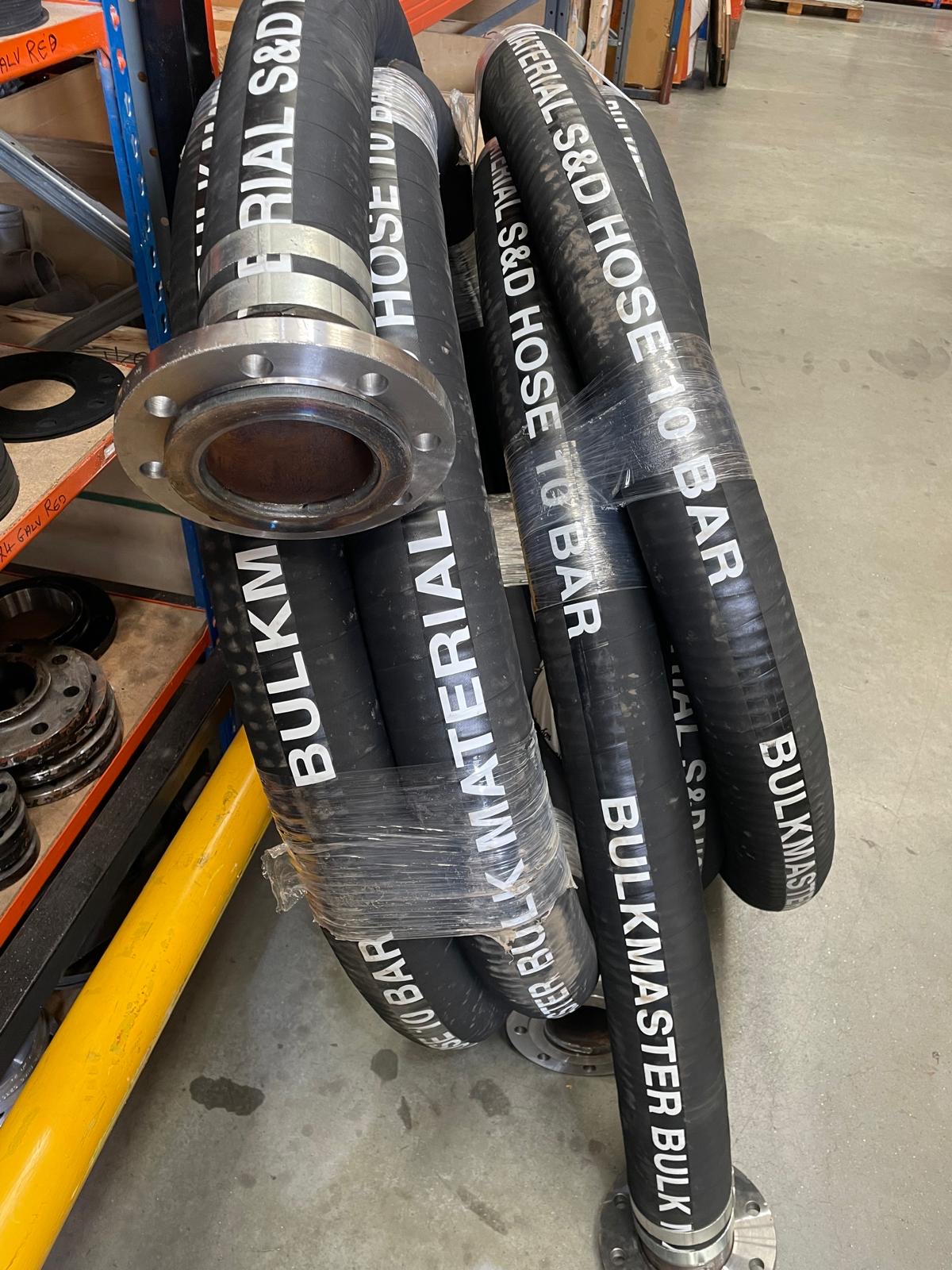 RUBBER HOSE ASS Large black industrial rubber hoses with metal fittings, coiled and wrapped in plastic on a warehouse floor.